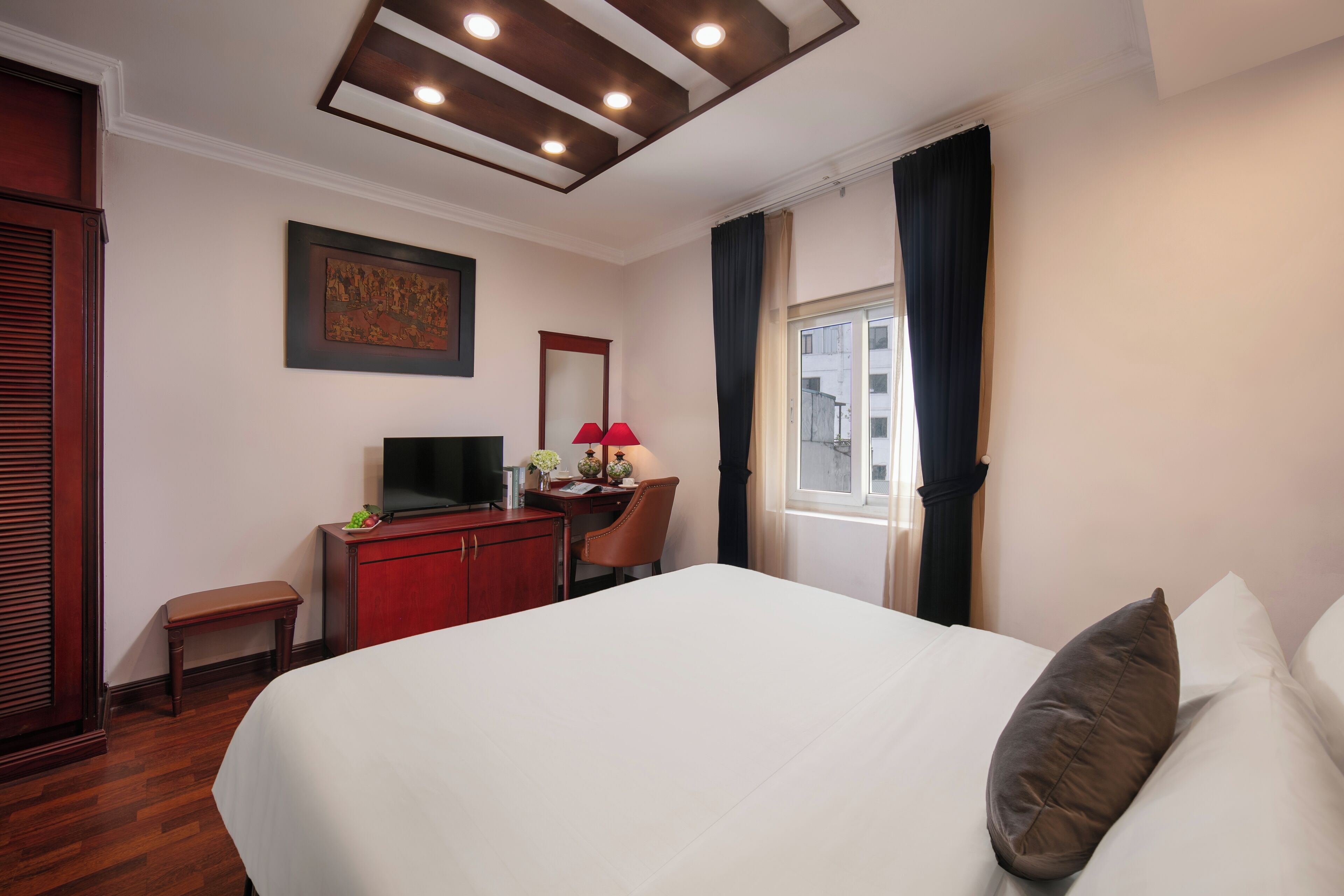 A hotel room with a large bed, a desk with a television, a small table, and a window with curtains.