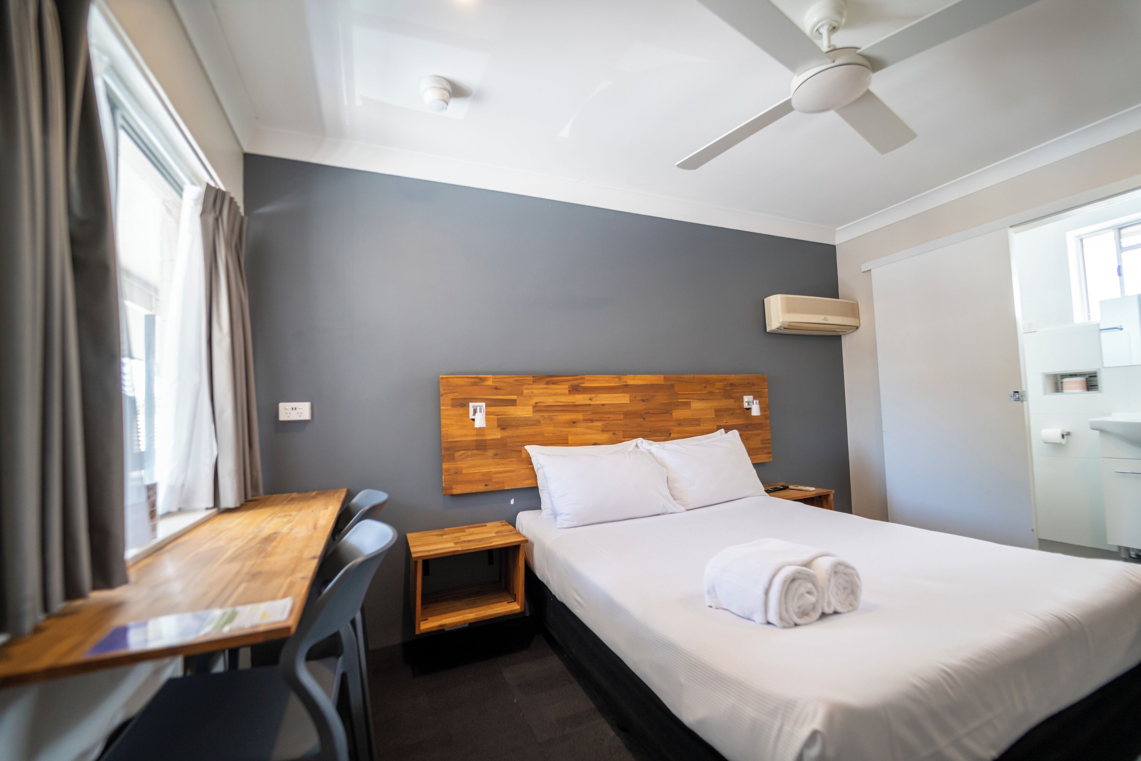Quarto de hotel com cabeceira de madeira, cama com roupa de cama branca, escrivaninha de madeira, uma cadeira, mesa de cabeceira pequena e um ventilador de teto.