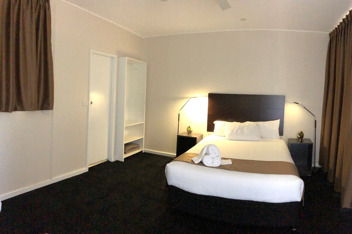 Quarto de hotel com cama, mesas de cabeceira, um criado-mudo, uma luminária e um guarda-roupa.