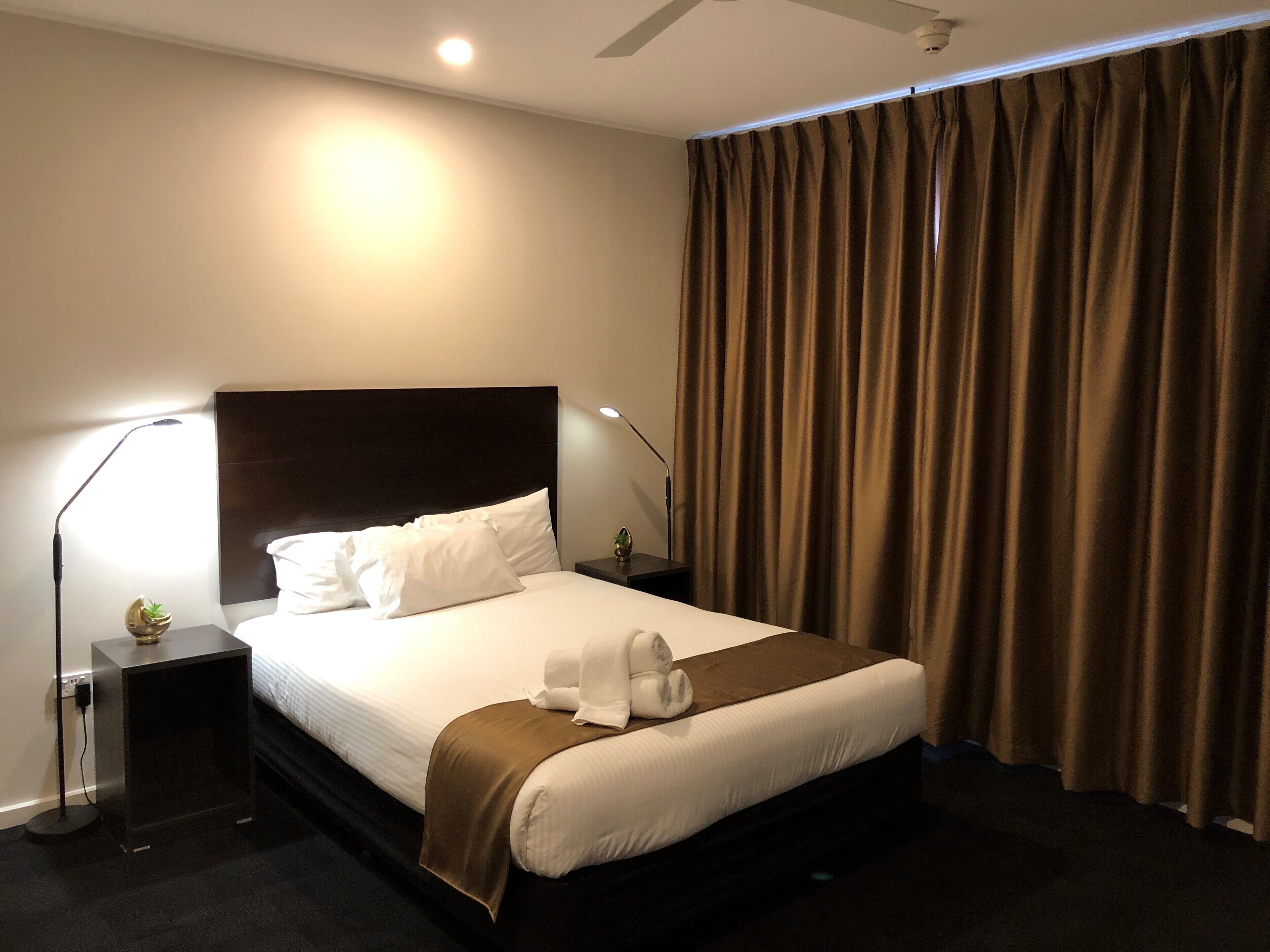 Una habitación de hotel con una cama grande, dos mesitas de noche, un ventilador de techo y una ventana con cortinas.