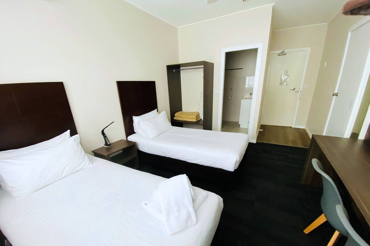 Quarto de hotel com duas camas de solteiro, uma escrivaninha, uma cadeira, um espelho e uma porta.