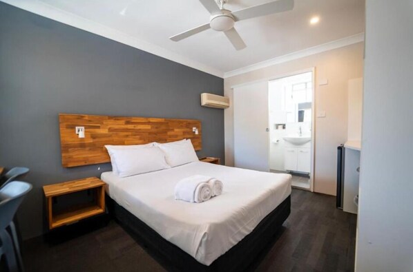 A hotel room with a wooden headboard, a bed with white linens, a ceiling fan, a bedside table, and a view of the bathroom.