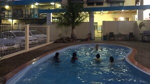 A swimming pool with four people in the water, surrounded by a fenced area with parked cars.