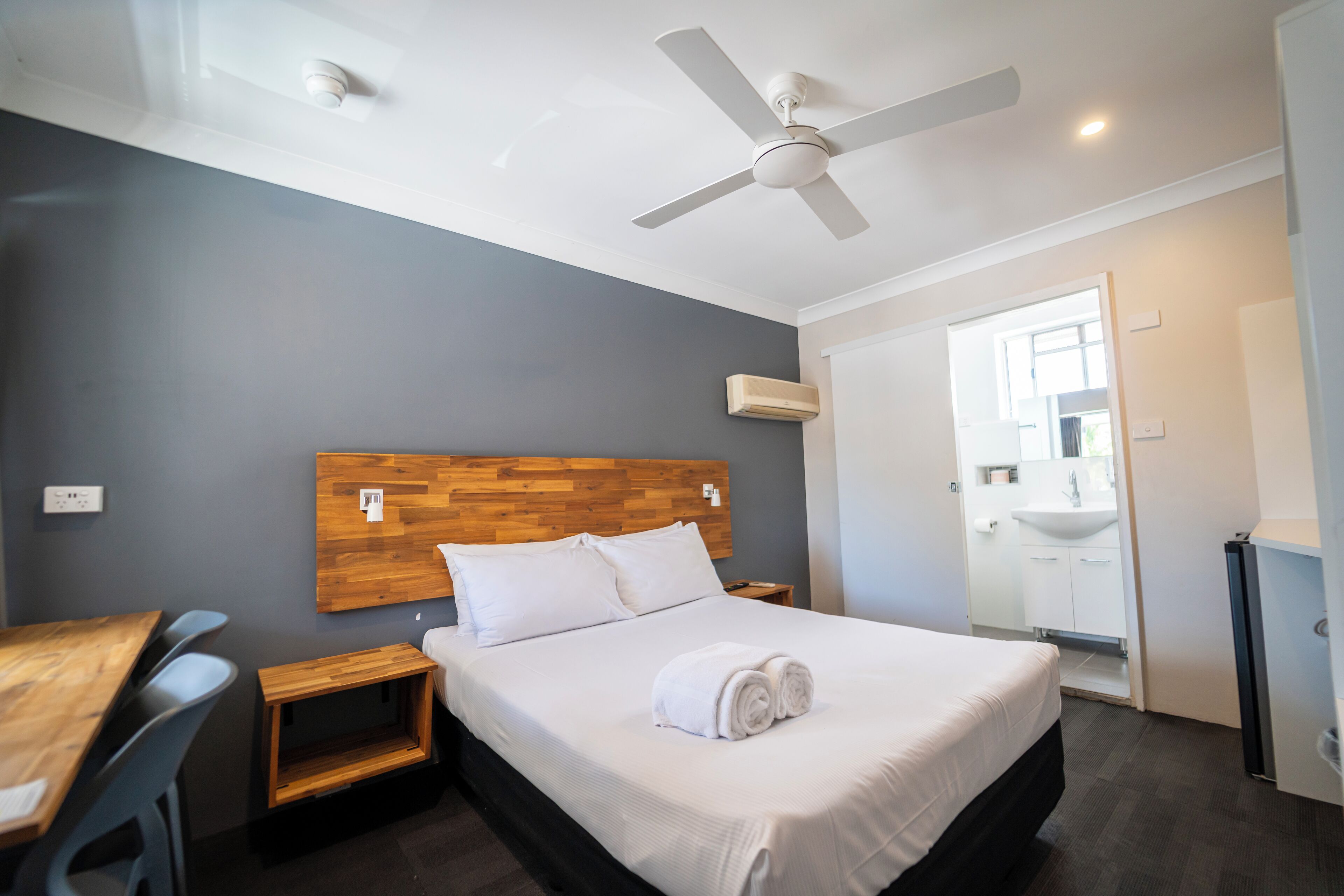 Hotelkamer met houten hoofdeinde, een bed met witte lakens, een bureau met stoelen, een plafondventilator en een badkamer met wastafel en spiegel.