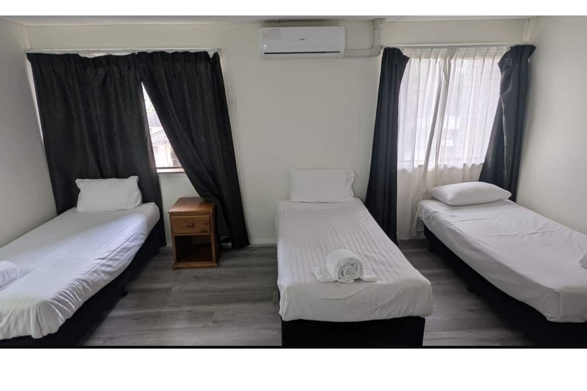 Quarto com três camas de solteiro, uma mesa de cabeceira de madeira e uma janela com cortinas.