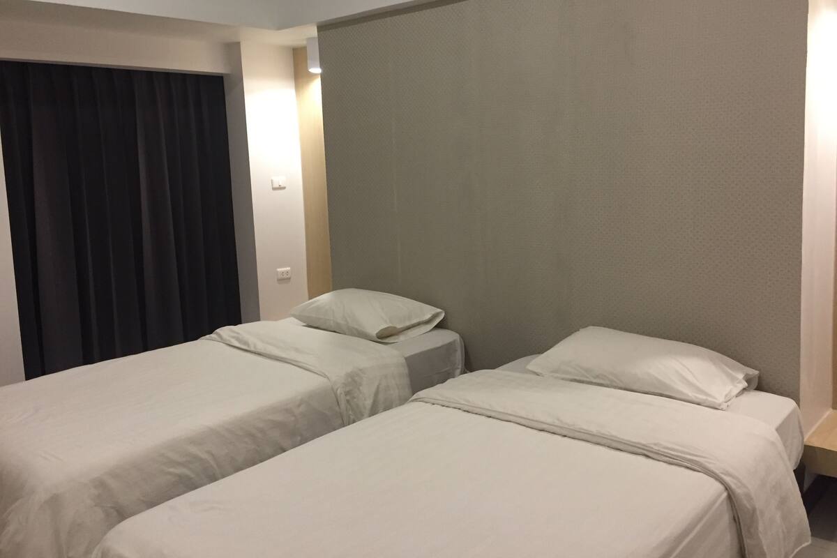 Una habitación de hotel con dos camas individuales, una cortina oscura, un interruptor de luz y una lámpara fijada a la pared.