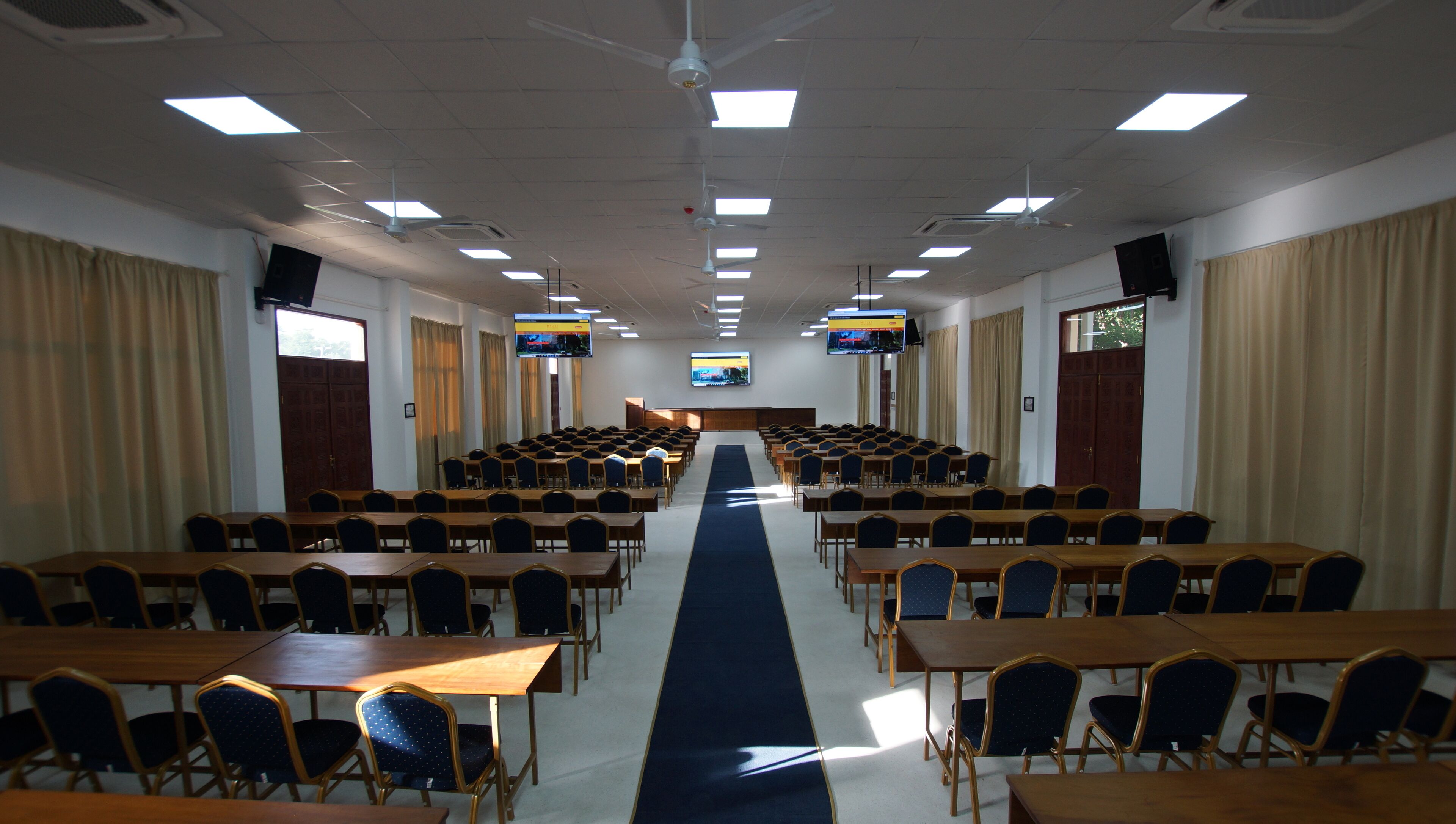 A conference room with rows of tables and chairs, a blue carpet, ceiling fans, and a large screen at the front.