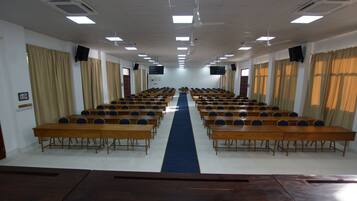 A conference room with rows of wooden tables and chairs, a blue carpet, and ceiling fans.