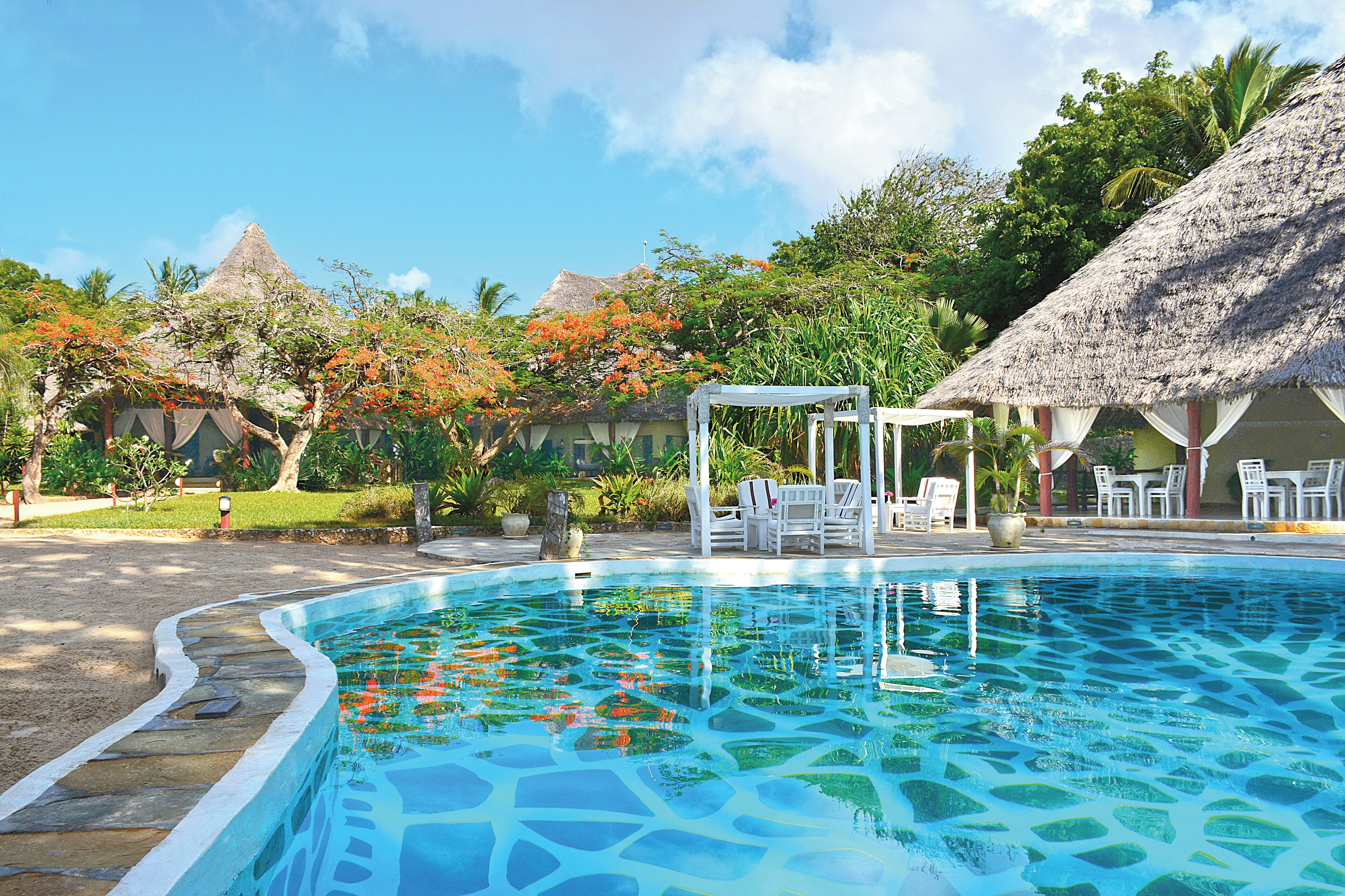 A swimming pool with a mosaic tile design, surrounded by thatched-roof structures and white outdoor furniture.