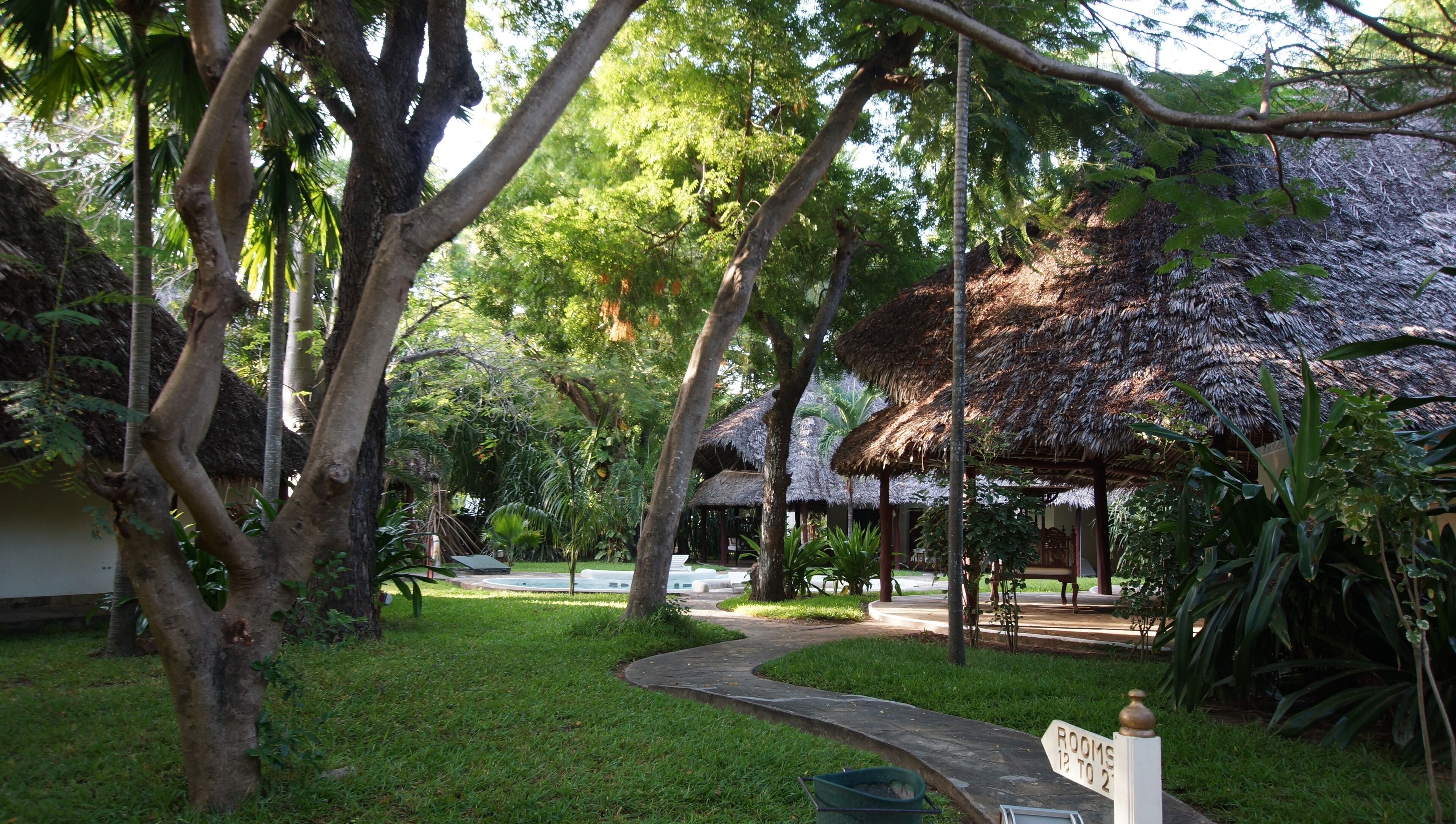 A garden pathway leading to a thatched-roof structure, surrounded by lush greenery and trees.