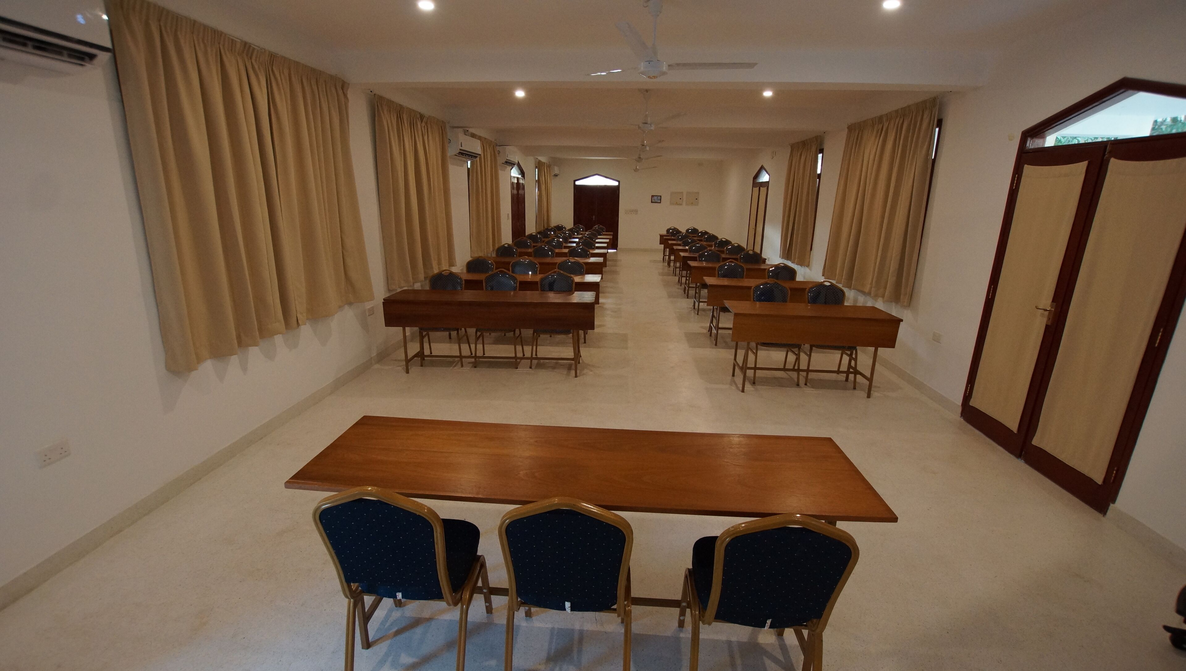 A conference room with long tables and chairs, beige curtains, and ceiling fans.