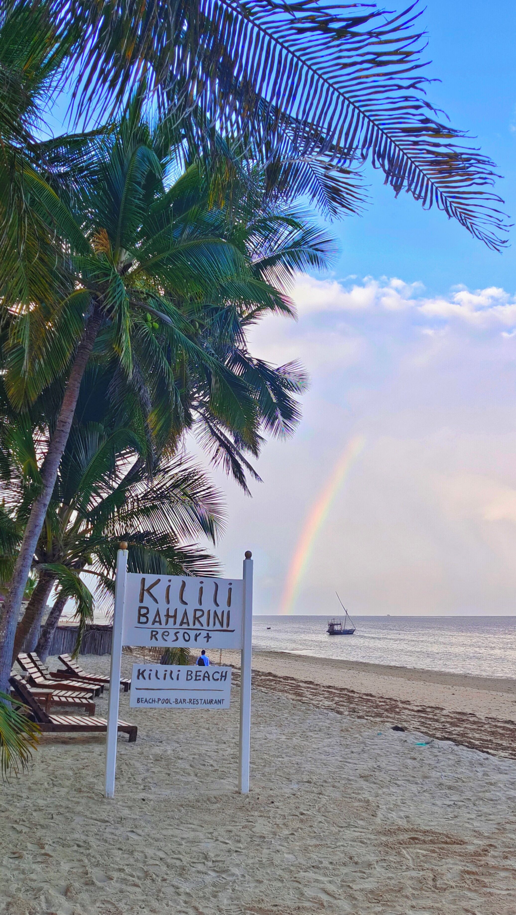 A beach sign for 'Kilili Baharini Resort' with palm trees and a rainbow in the background.