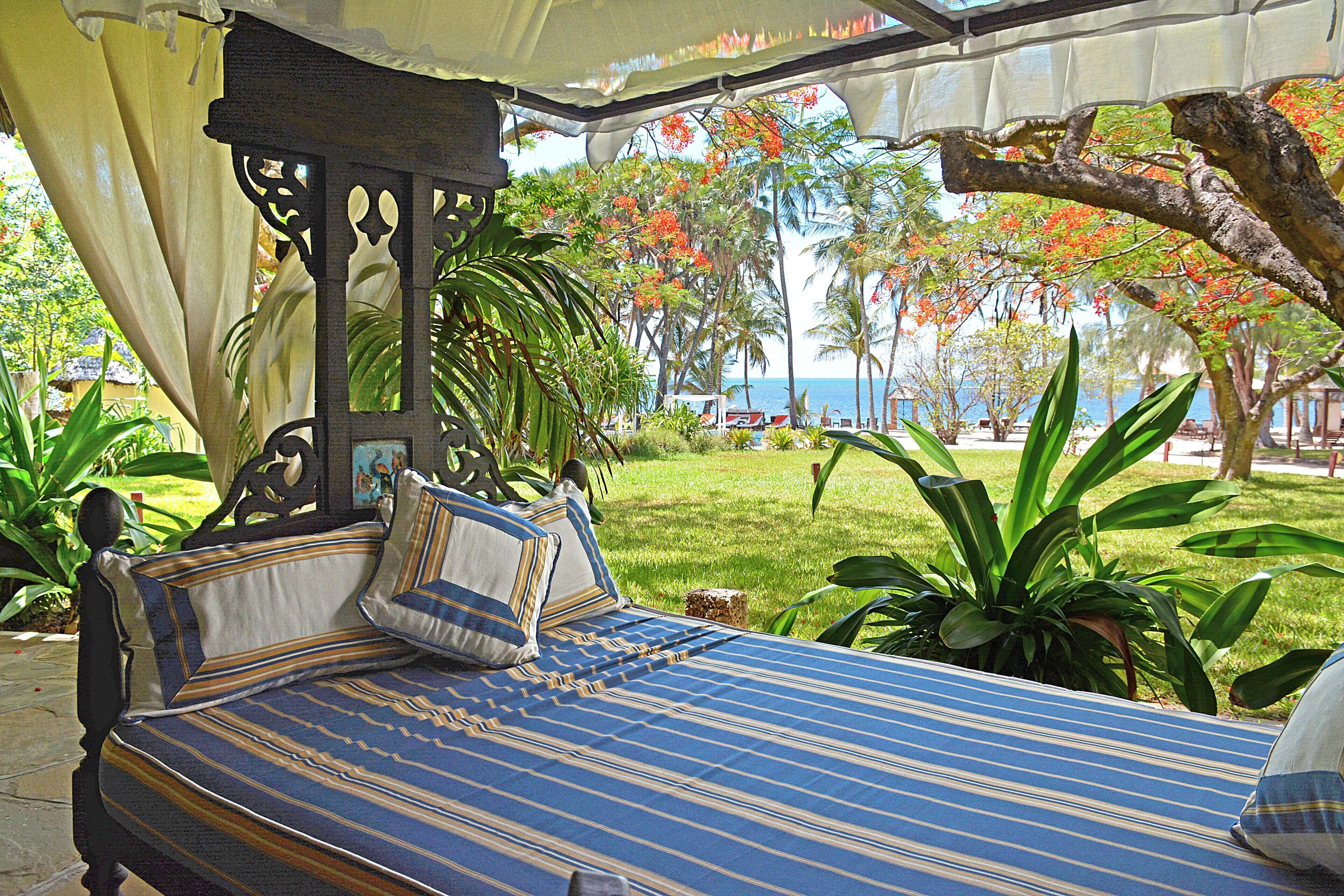 A bed with a striped blue and white bedspread, a canopy overhead, and a view of a tropical garden with palm trees and a beach in the background.