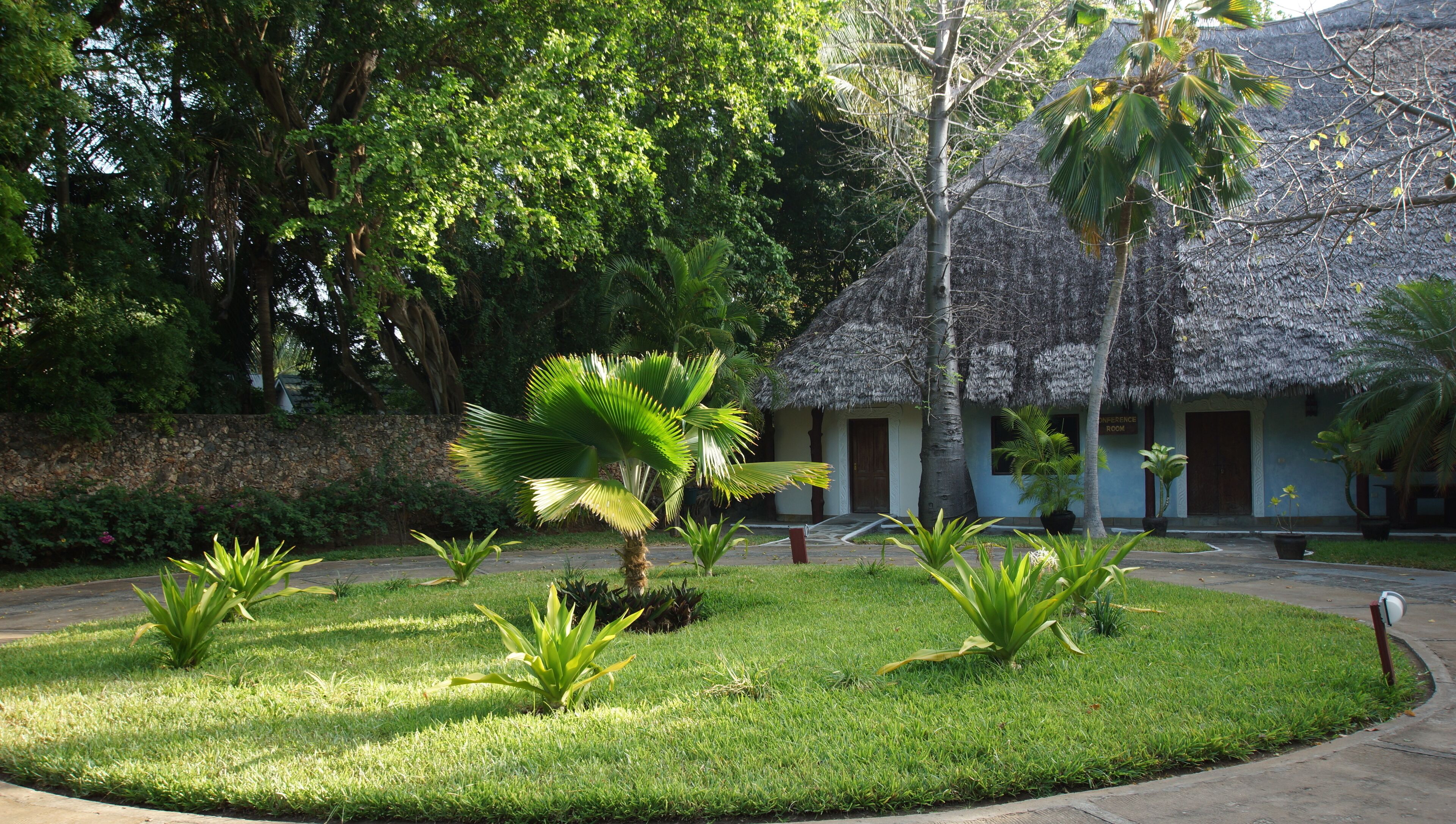 A well-maintained garden with a thatched-roof building, surrounded by lush greenery and a stone wall.