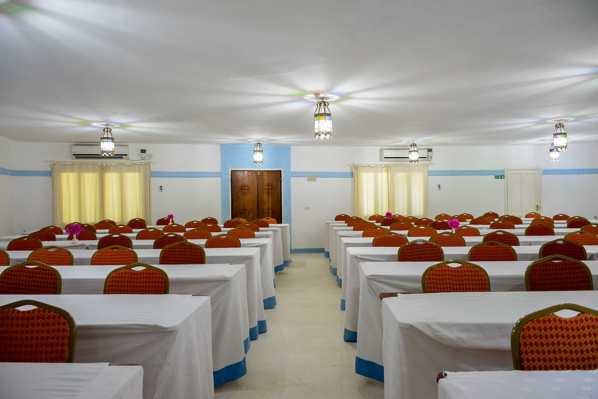 A conference room with rows of tables and chairs, air conditioning units, and ceiling lights.