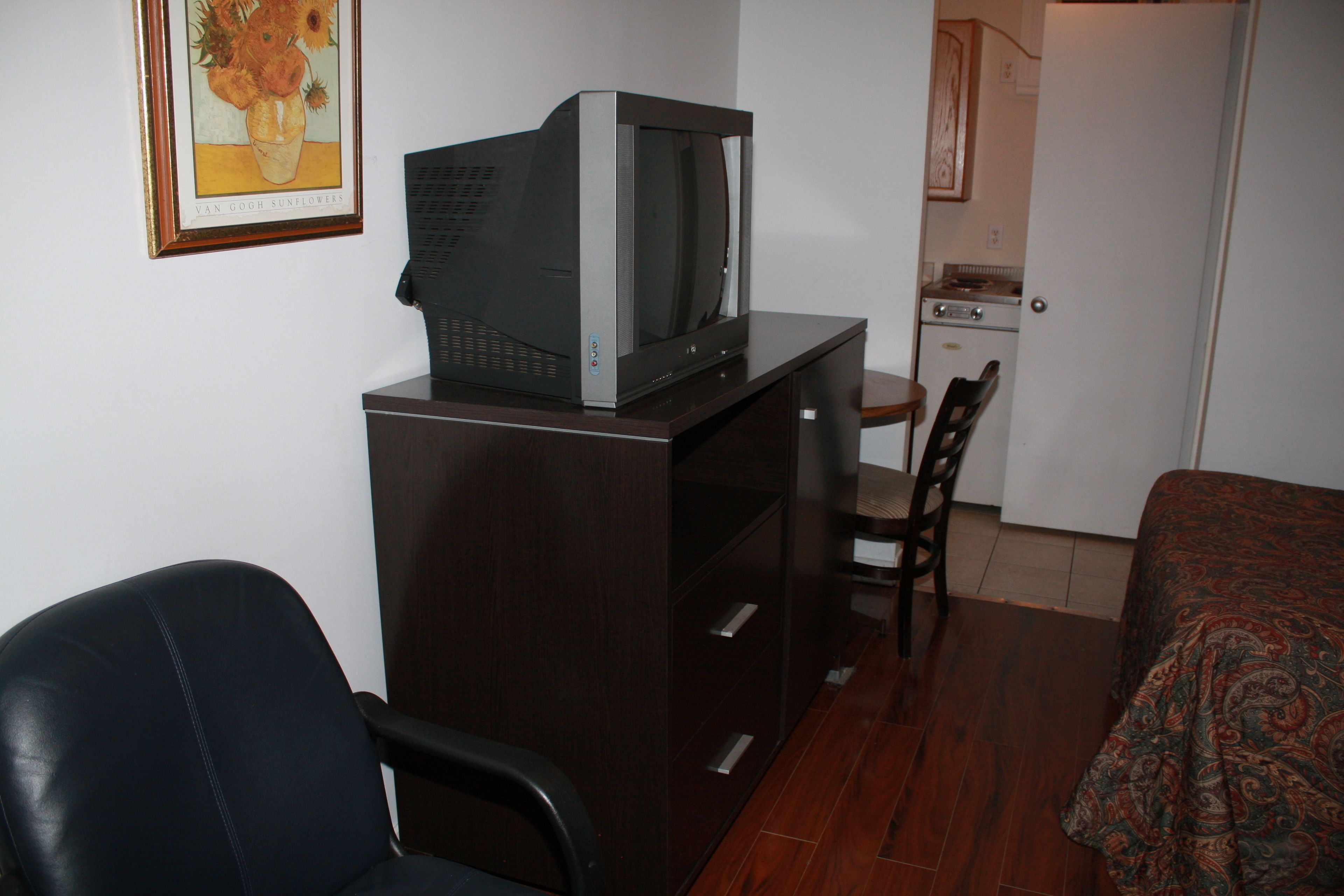 A compact room with a bed, a desk with a television, a chair, a small kitchenette, and a door leading to another room.