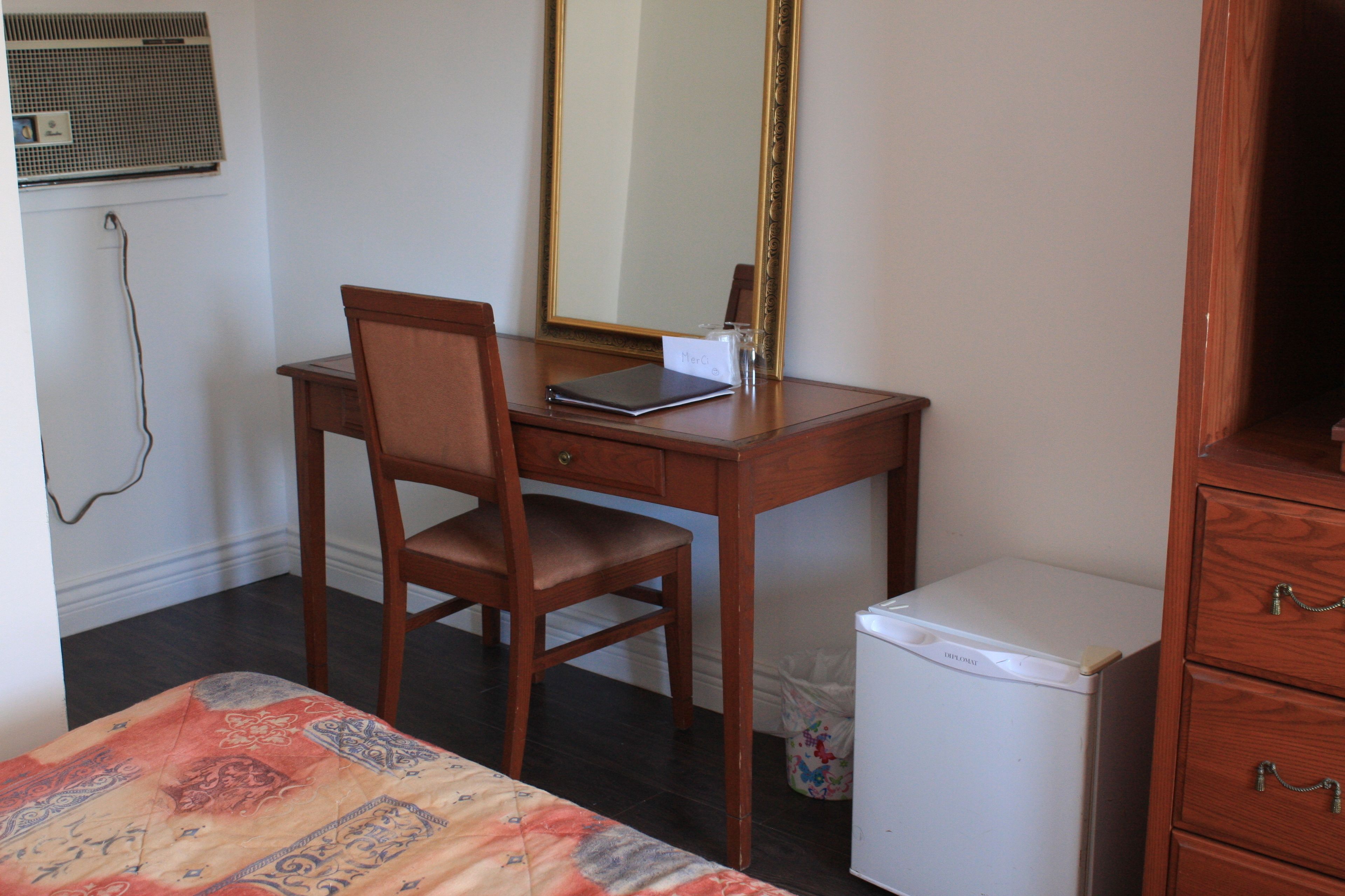 A small, neatly arranged bedroom with a wooden desk, a chair, a bed with a patterned bedspread, a white refrigerator, and a wooden dresser.