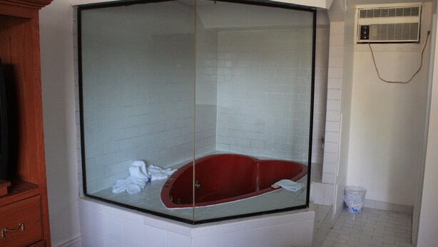 A corner bathroom with a glass-enclosed bathtub, white tiled walls, and a wooden cabinet. - Grand Motel Saint-Hubert (Longueuil)