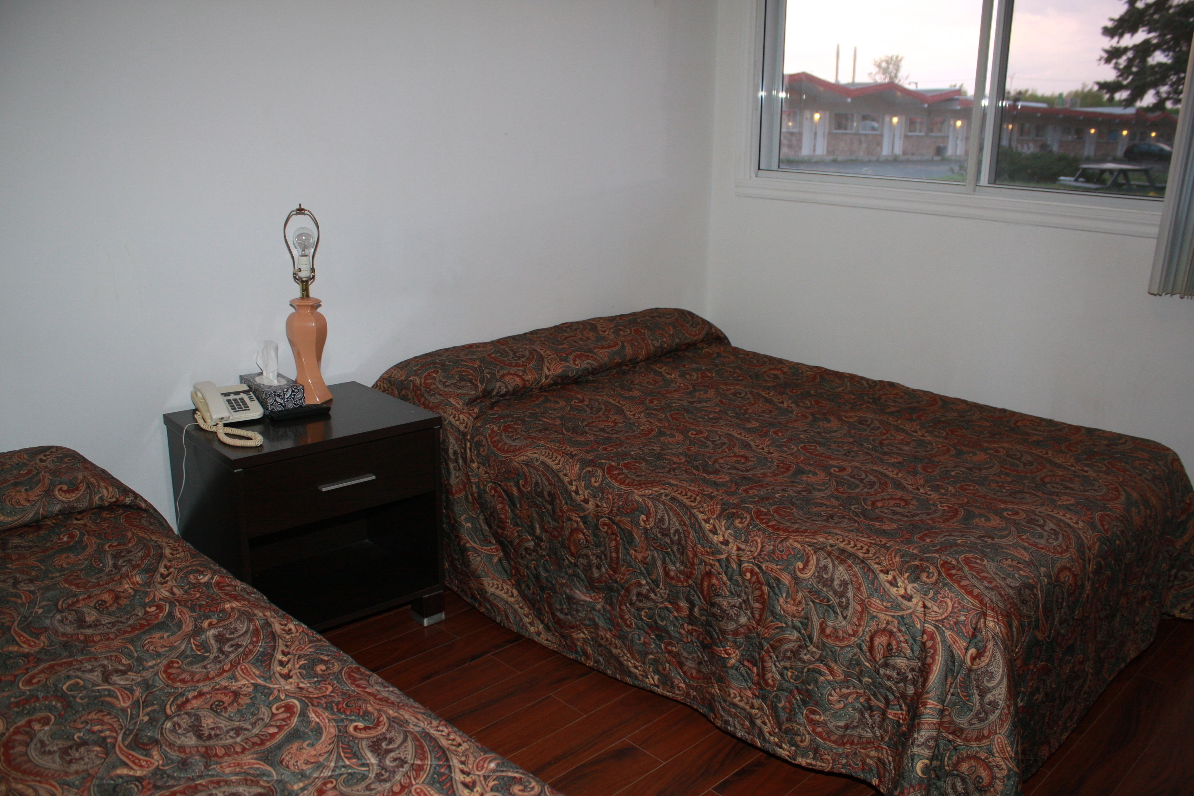 A room with two beds, a nightstand with a lamp, and a window with a view of buildings.