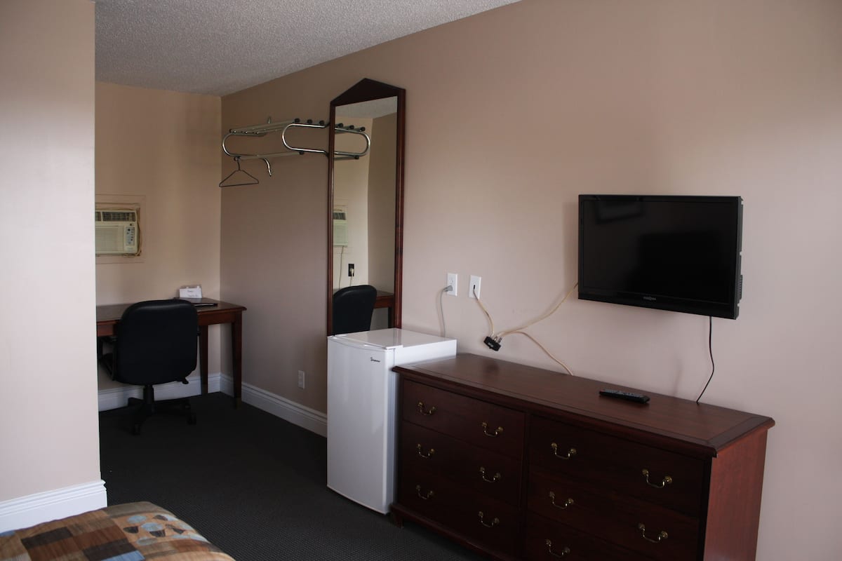 Una habitación de hotel con cama, escritorio con silla, cómoda, espejo, televisión y refrigerador.