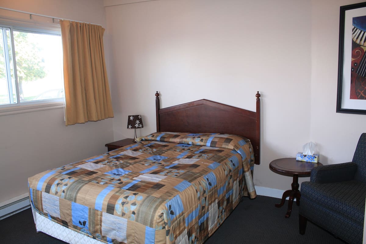 Una cama bien hecha con una colcha estampada, una mesita de noche con una lámpara, y una silla en una habitación con ventana y un cuadro en la pared.