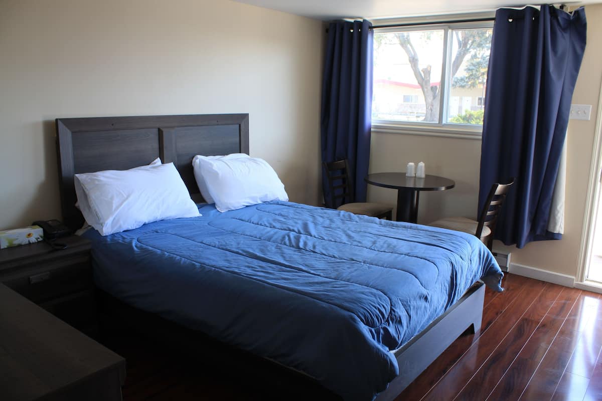 Una cama bien hecha con colcha azul y almohadas blancas, una mesita con dos tazas, y una silla junto a la ventana con cortinas oscuras.