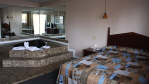 A hotel room with a large bed, a wooden headboard, a small bedside table, a nightstand, a mirror, a bathroom with a bathtub, and a towel rack. - Grand Motel Saint-Hubert (Longueuil)