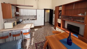Cocina compacta con armarios blancos, microondas y lavabo. Zona de comedor con mesa y sillas, y estantería con libros y objetos decorativos.