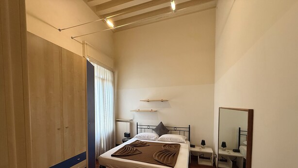 Un dormitorio con cama, mesitas de noche, un espejo, un armario y una ventana con cortinas.