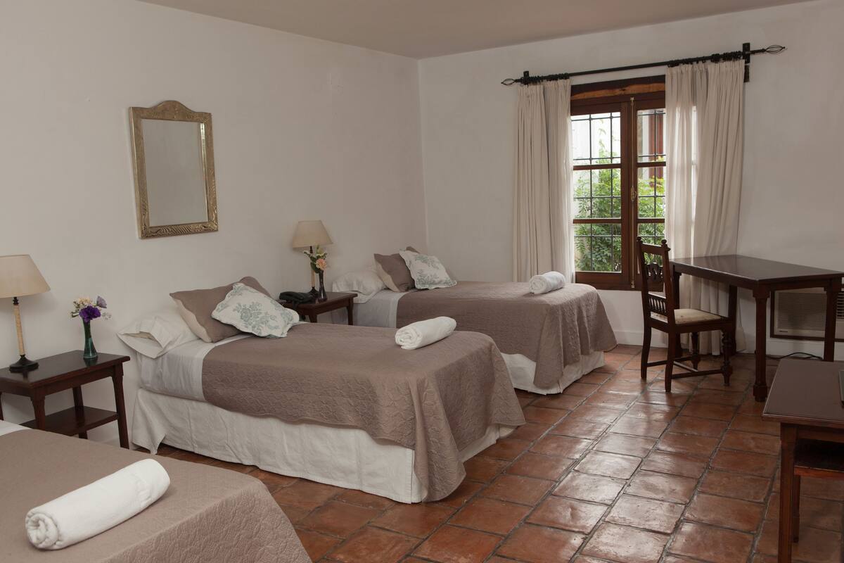 A hotel room with two beds, a table, a chair, a mirror, and a window with curtains.
