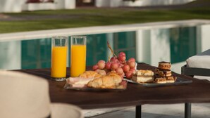 Une table de petit-déjeuner avec des croissants, des raisins et des verres de jus, placée devant une piscine et offrant une vue sur un bâtiment blanc aux grandes fenêtres, avec un lampadaire à proximité.