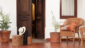 Une porte en bois ornée de sculptures complexes, une chaise en osier avec un coussin et deux plantes en pot sur un sol carrelé.
