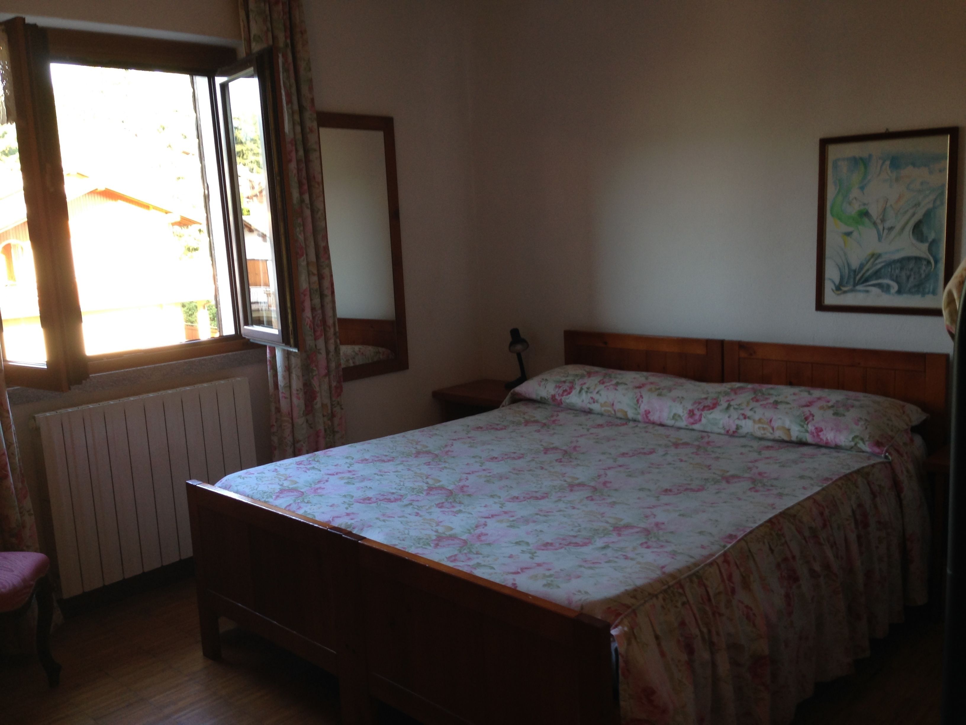 Una habitació amb un llit de fusta, una tauleta de nit, una cadira, un radiador, una finestra amb cortines i un quadre a la paret.