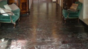 A spacious hallway with a wooden ceiling and marble flooring, flanked by green upholstered chairs and a wooden cabinet.
