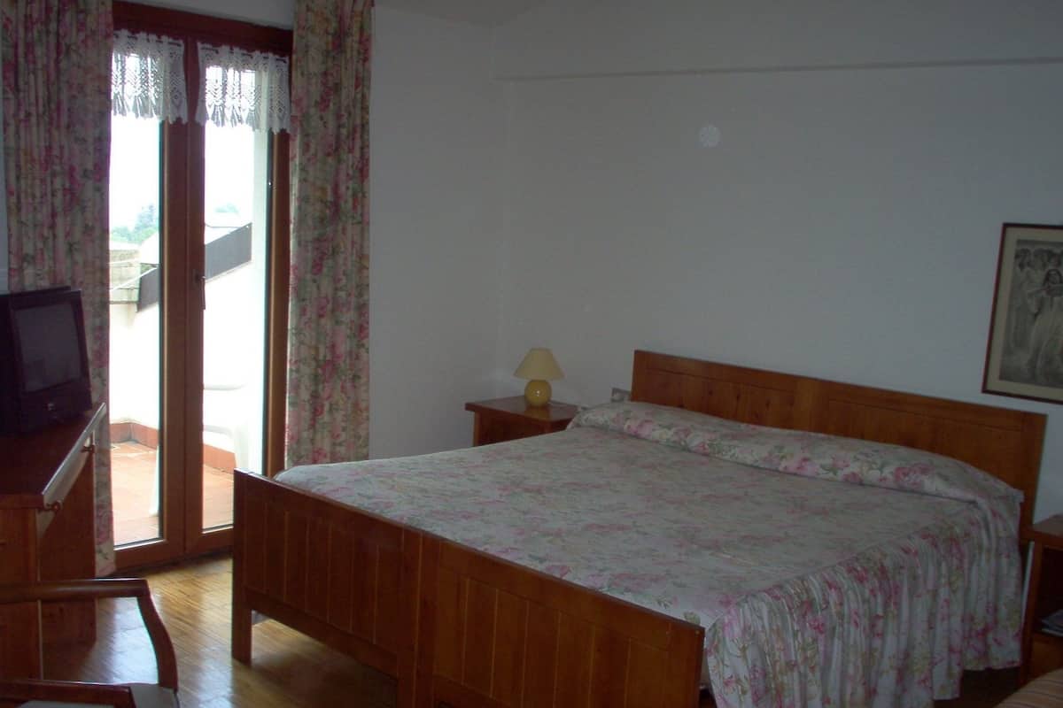 A bedroom with a wooden bed, a desk with a television, a nightstand with a lamp, and a view through a glass door.