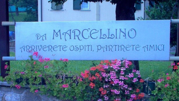 A sign with the text "DA MARCELLINO" and "ARRIVERETE OSPITI, PARTIRETE AMICI" in front of a stone wall with flowering plants.