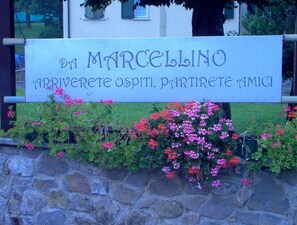 A sign with the text "DA MARCELLINO" and "ARRIVERETE OSPITI, PARTIRETE AMICI" in front of a stone wall with flowering plants. - Hotel Marcellino (Selvino)