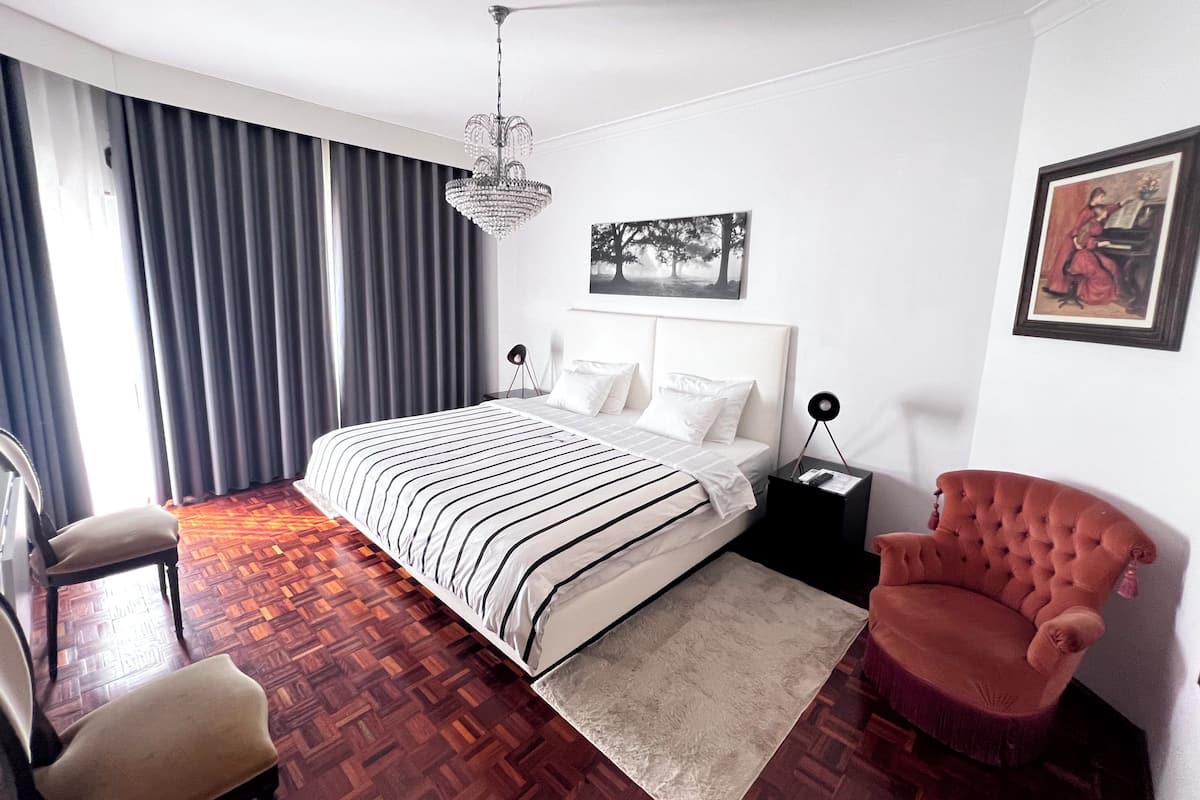 Une chambre d’hôtel moderne avec un grand lit, un fauteuil rouge, un parquet en bois et un lustre.