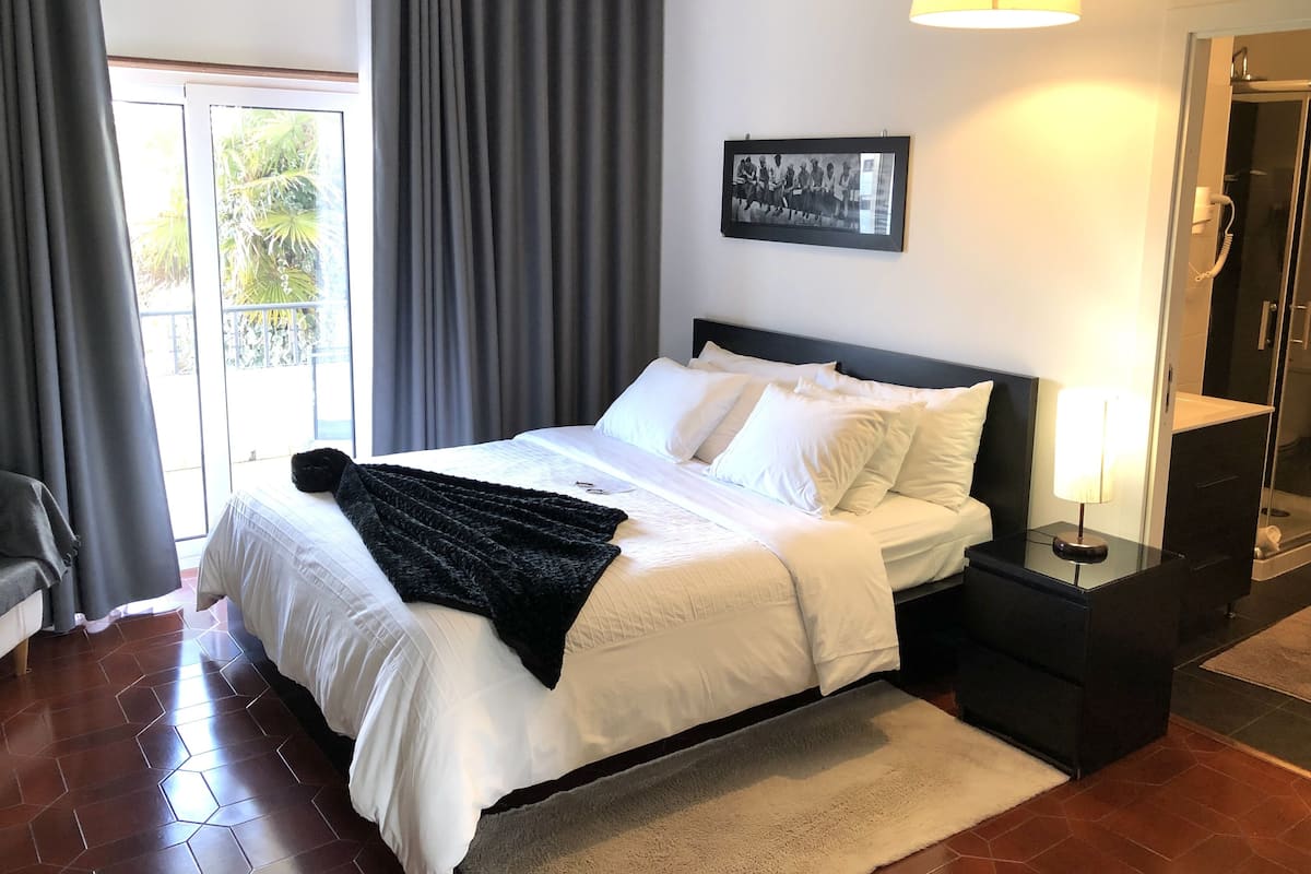 Une chambre d’hôtel avec un grand lit, une table de chevet avec une lampe, une photo encadrée accrochée au mur et une salle de bain visible à travers une porte ouverte.