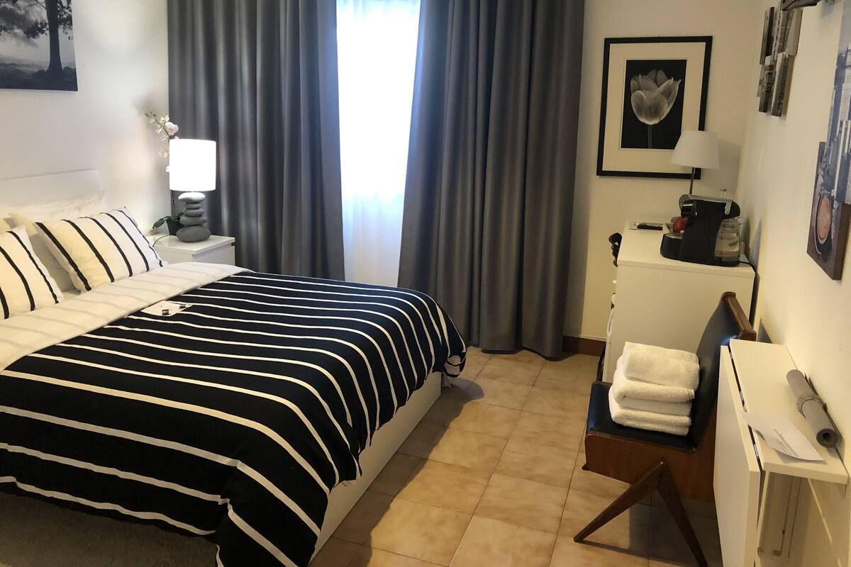 Une chambre d’hôtel bien aménagée, avec un lit, une table de chevet dotée d’une lampe, un petit bureau avec une machine à café et une chaise.