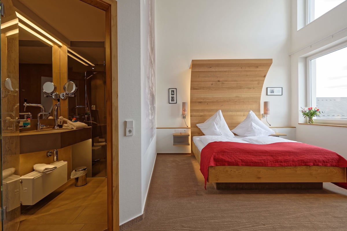 A modern hotel room with a wooden headboard, a large mirror, a built-in vanity, and a red bedspread.