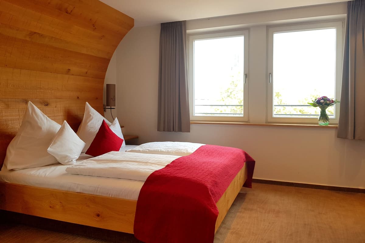 A modern bedroom with a wooden headboard, a large bed with white and red bedding, a nightstand, and a window with curtains.