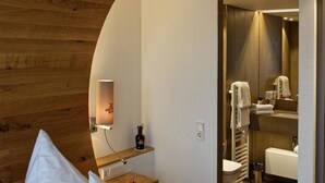 Ein Hotelzimmer mit hölzernem Kopfteil, einem Bett mit weißer Bettwäsche, einem roten Akzent, einem Nachttisch mit einer Flasche, einem Spiegel und einem durchgegangene Tür erkennbaren Badezimmer.