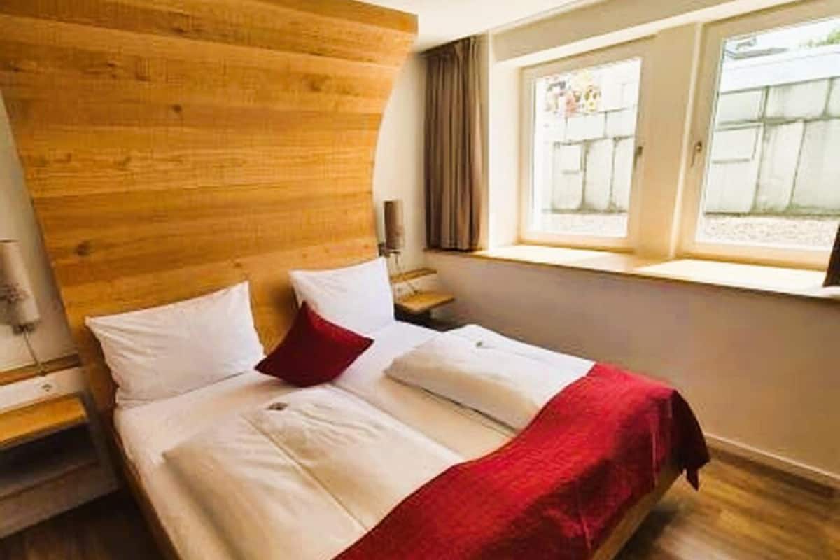 A hotel room with a wooden headboard, a bed with white linens and a red throw, a nightstand, and a window with curtains.