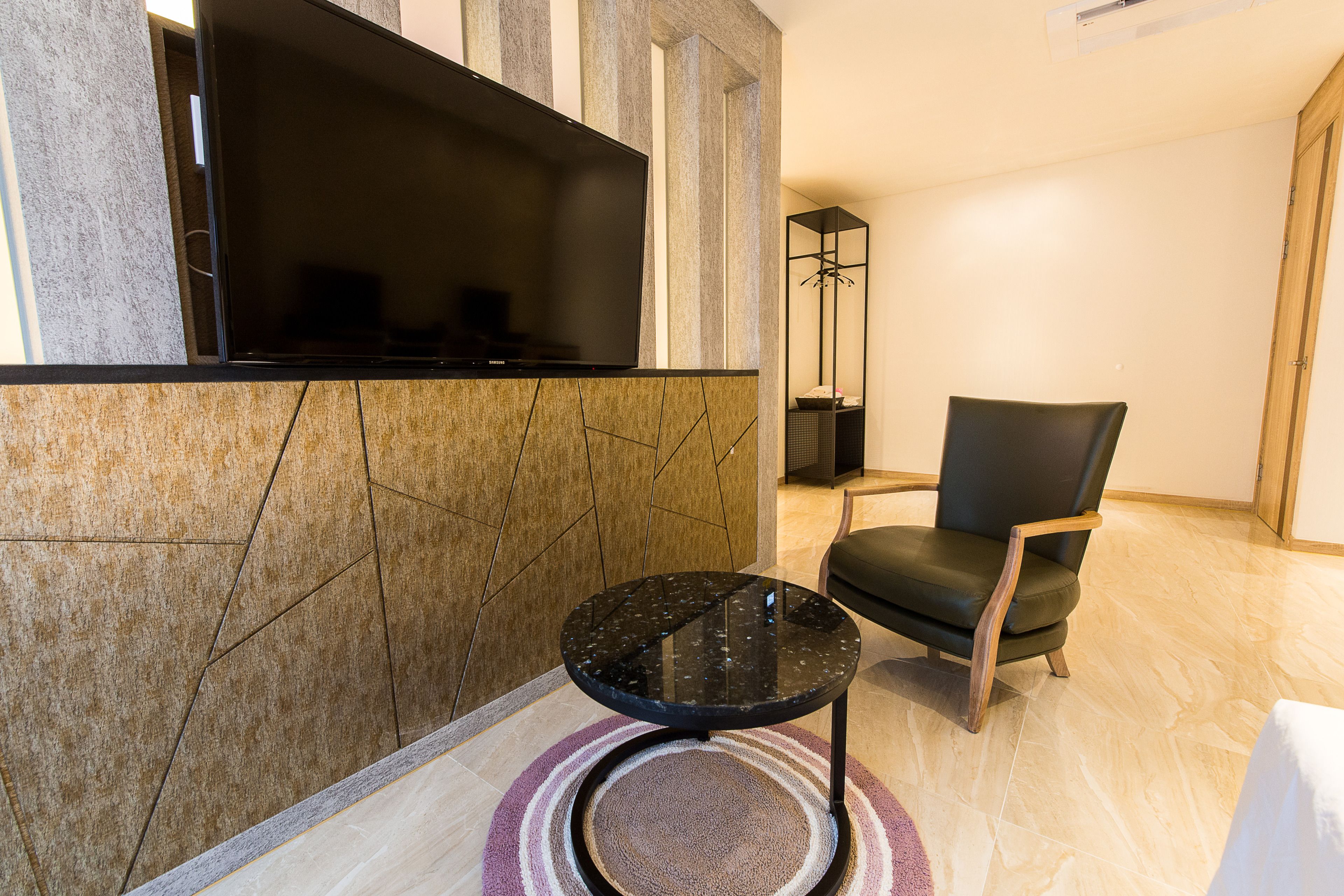 A modern living room with a flat-screen TV mounted on a textured wall, a round black coffee table, a leather armchair, and a floor lamp.