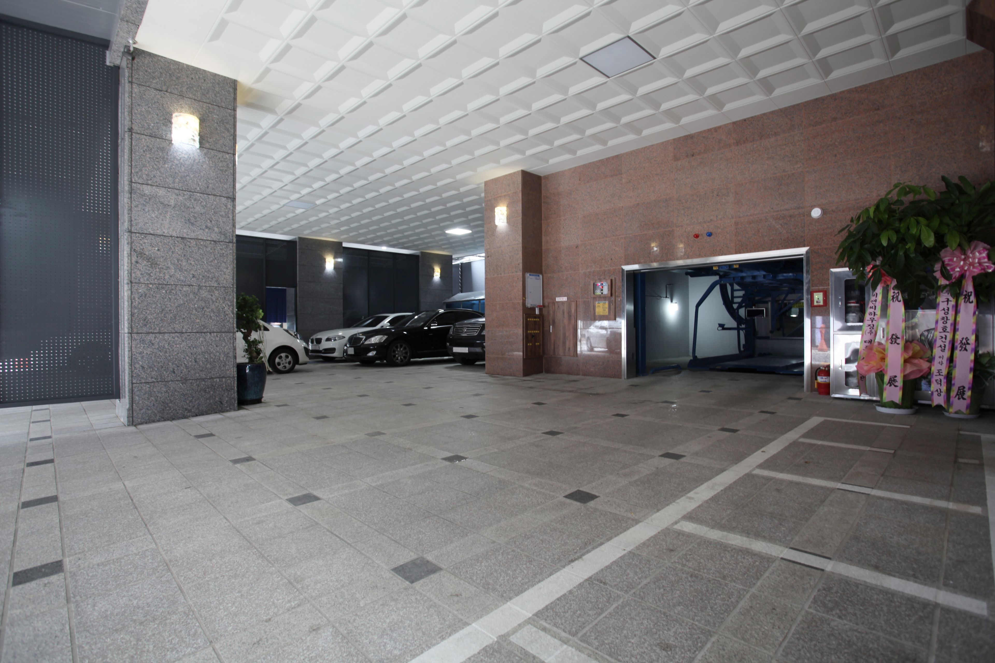 A spacious lobby with a tiled floor, a parking area with several cars, and a reception desk with a plant.