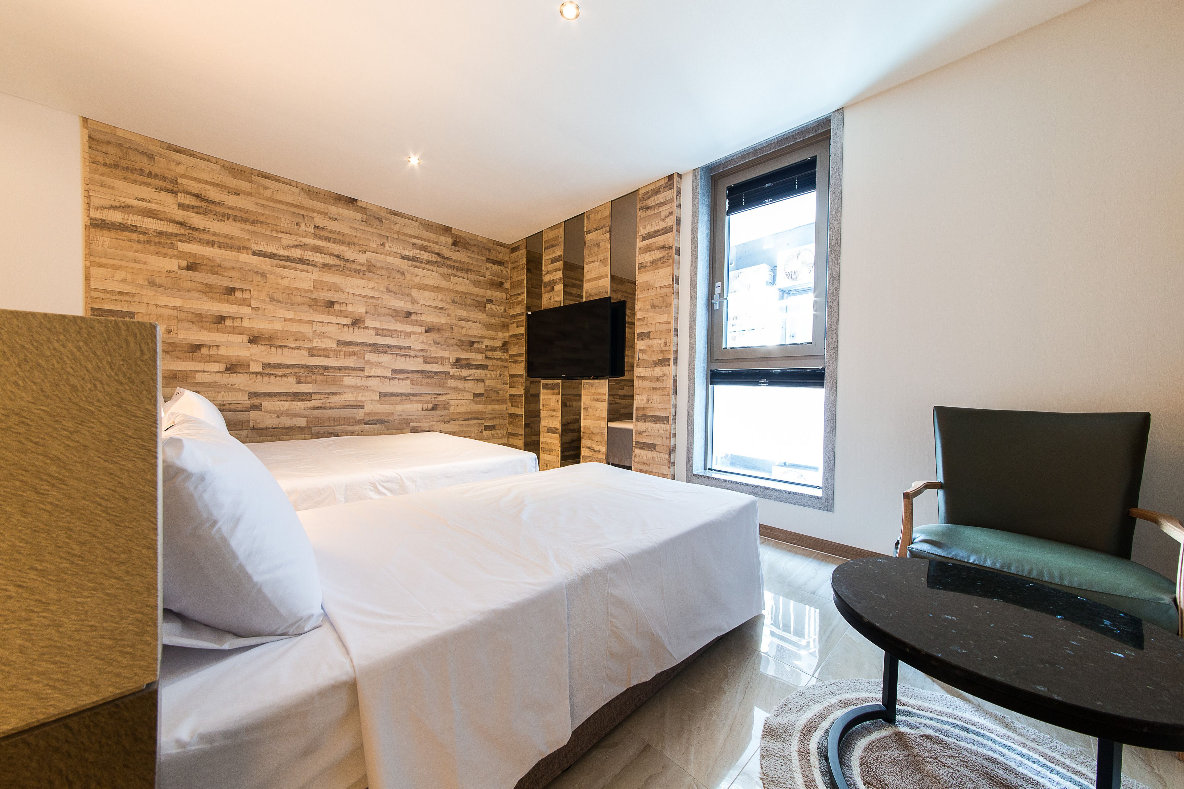 A modern hotel room with two beds, a flat-screen TV, a green armchair, and a large window.