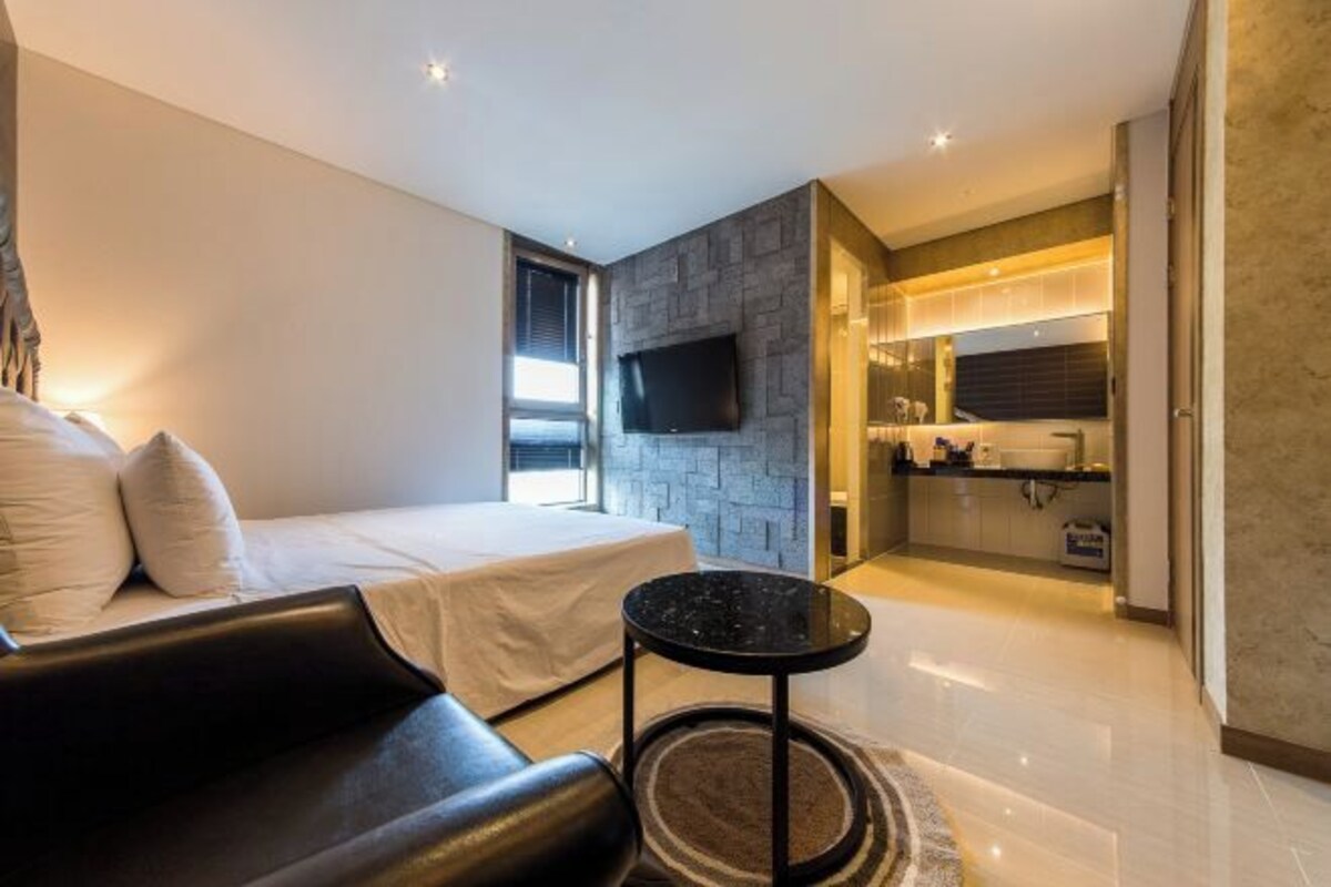 Quarto de hotel moderno com uma cama, um sofá de couro preto, uma pequena mesa redonda e uma televisão fixada na parede.