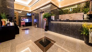 Een modern ingerichte hotelreceptie met een receptiebalie, een digitaal scherm en een plantenarrangement.