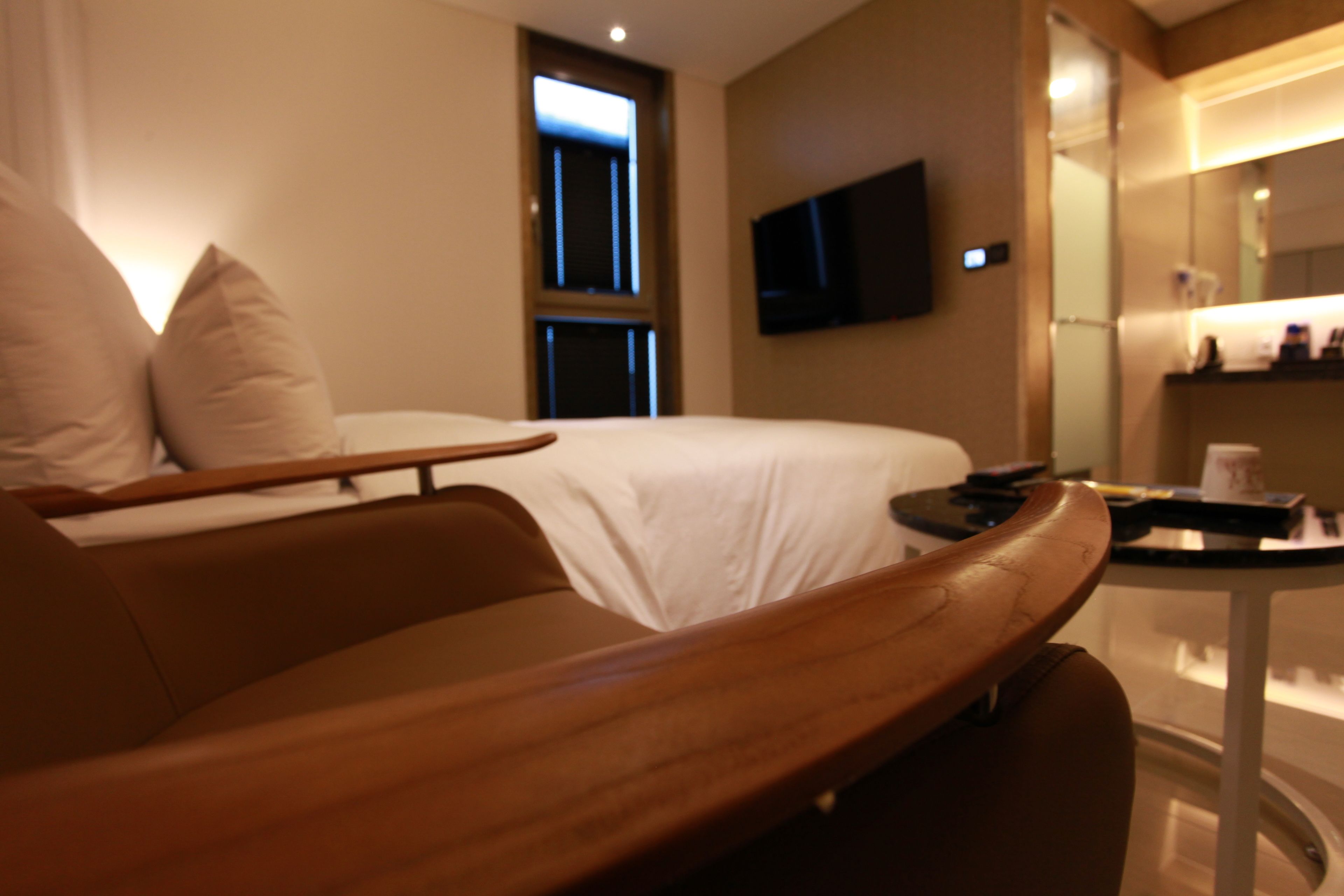 A hotel room with a bed, a wooden chair, a small table, a TV mounted on the wall, and a window.